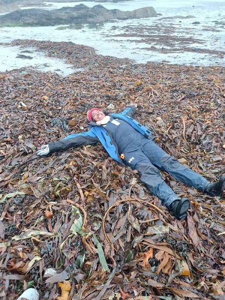 Priya lying on top of kelp on the seashore dressed in blue overalls spread out like a starfish